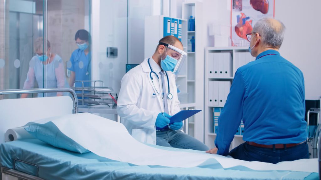 Médico com EPIs conversando com um paciente que está sentado em uma maca no consultório.