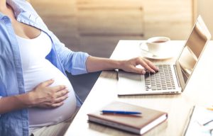 Mulher grávida sentada à mesa usando um notebook com uma mão enquanto segura a barriga com a outra mão.